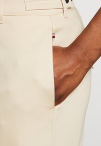 Pantalones beige de material suave de algodón, con un bolsillo lateral y un pequeño logo de la marca en rojo, blanco y azul en la costura del bolsillo.