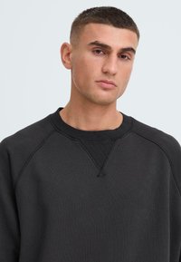 Felpa nera con texture a coste, maniche raglan e scollatura a girocollo. Presenta un piccolo dettaglio cucito a triangolo all'altezza del collo.