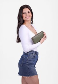 Green zip wallet held against a fitted white top, paired with a high-waisted denim skirt. Smooth texture, simple design, no visible patterns.