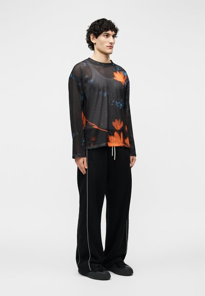 Homme portant un haut à manches longues en maille noire avec imprimé floral orange, pantalon noir ample avec liseré blanc et baskets noires, debout devant un fond blanc.