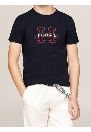Chłopiec nosi granatowy T-shirt z czerwonym konturem litery "H" i napisem "Hilfiger", zestawiony z białymi spodniami i markowym paskiem.