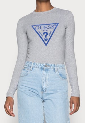 Person trägt ein graues Langarmshirt mit blauem GUESS-Logo und hochtaillierte hellblaue Jeans.