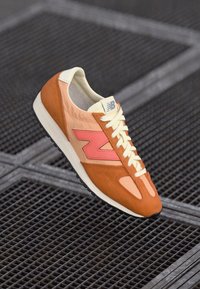 Sneaker New Balance avec un dessus orange et rose, en matériaux en daim et en maille, des lacets crème et une semelle texturée. Détails de logo notables présents.