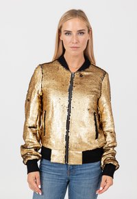 Donna in piedi che indossa una giacca bomber dorata con paillettes, polsini e vita neri, abbinata a jeans blu su uno sfondo chiaro e semplice.