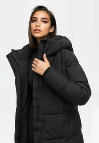 Chaqueta de plumas negra con capucha, cierres de cremallera y a presión, textura acolchada y un logo en la manga. Material suave e insulated.
