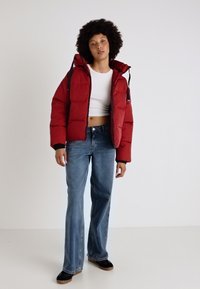 Chaqueta acolchada roja con capucha, con un diseño acolchado y detalles negros. Combinada con jeans acampanados de color azul claro y zapatos negros sin cordones.