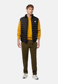 Gilet noir rembourré avec col montant, porté sur un sweat-shirt jaune, associé à un pantalon olive et des baskets blanches.