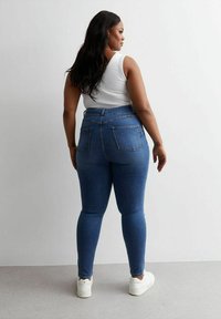 Jeans ajustados en denim azul oscuro con una cintura alta, que cuentan con dos bolsillos traseros y un ajuste ceñido. Combinados con una camiseta sin mangas blanca y zapatillas blancas.