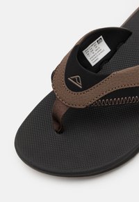 Brun syntetisk flip-flop med teksturert svart såle. Har en vevd tåpost og sømdetaljer. Merket er synlig på innersålen.