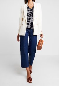 Blazer blanco de doble botonadura sobre una camiseta de cuello en V a rayas blancas y negras, pantalones vaqueros de pierna ancha azul oscuro y un bolso marrón texturizado. Chanclas marrones.