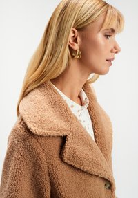 Blondynka w profilu, nosząca złote kolczyki w kształcie kół, białą koszulę w brązowe kropki oraz fakturowany beżowy płaszcz z szerokim kołnierzem.