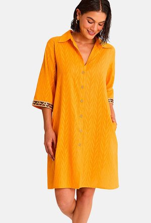 Femme portant une robe boutonnée orange vif avec des manches trois-quarts et des poignets à motifs, debout avec une main dans la poche, souriante.