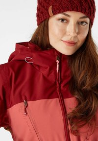 Rode en roze waterdichte jacket met een ritssluiting aan de voorkant, verstelbare capuchon en logo-detail, gecombineerd met een gebreide bordeauxrode beanie.