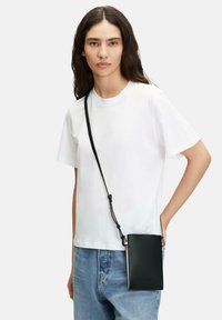 Black leather crossbody bag with a textured surface, rectangular shape, and adjustable strap, worn over a white t-shirt with blue jeans.
