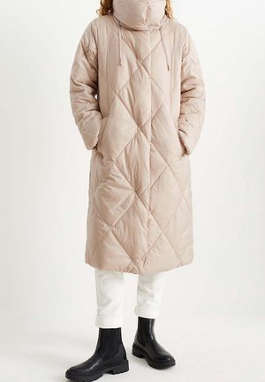 Persoon die een beige doorgestikte lange donzen jas draagt met hoge kraag en capuchon, witte opgerolde broek en zwarte stevige laarzen, staand.