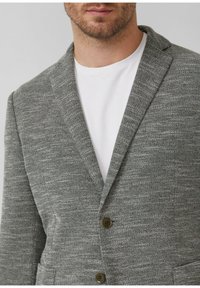 Grijze blazer gemaakt van textuurstof, met een notch lapel, twee voorzakken en bruine knopen. Draagt over een wit t-shirt.