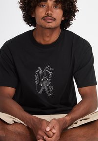 Hombre con cabello rizado que lleva una camiseta negra con arte lineal abstracto en blanco de figuras, flores y formas, sentado con las manos entrelazadas.