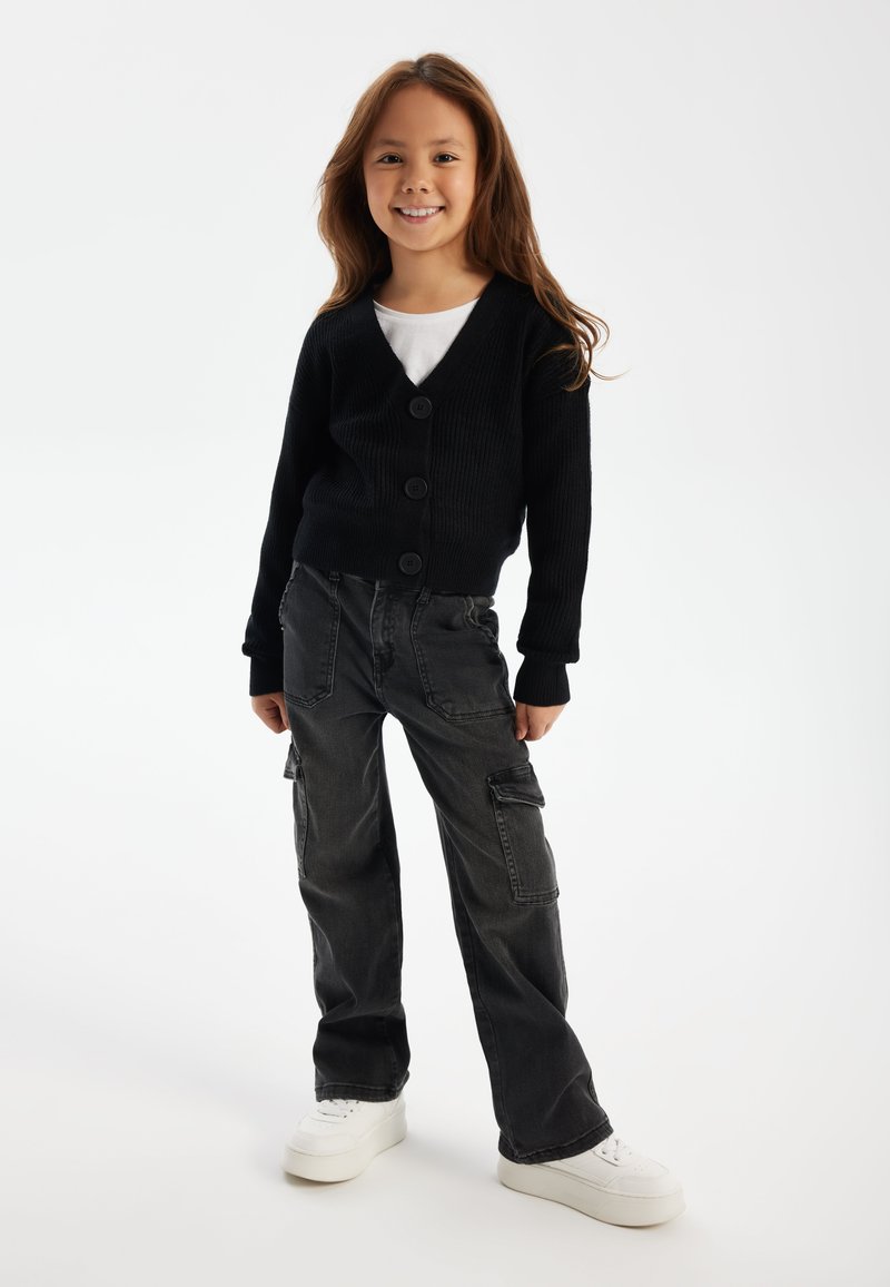 Cardigan noir côtelé à quatre boutons, porté sur une chemise blanche. Associé à un pantalon cargo gris foncé et des baskets blanches.