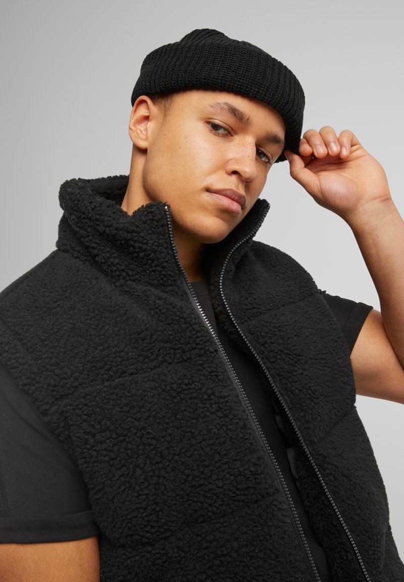 Black fleece vest with high collar and zip closure, featuring a textured surface. Worn over a black T-shirt, paired with a knitted black beanie.