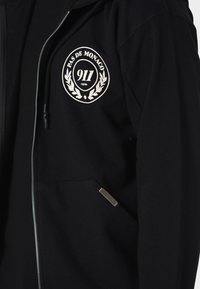 Black zip-up hoodie with white "Pas de Monaco 911 Club" emblem on chest and small rectangular label on pocket.