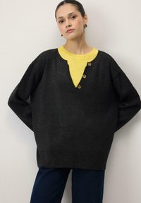 Pull noir surdimensionné avec un col en V, doté de trois boutons en bois et d'une chemise jaune contrastante en dessous.