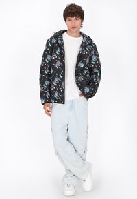 Jacket puffer negru cu imprimeuri colorate inspirate de spațiu, cu închidere tip fermoar și glugă, purtat cu pantaloni cargo bleu deschis și adidași albi.