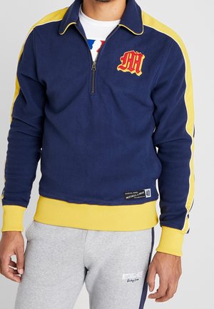 Homme portant un sweat-shirt bleu marine et jaune à col zippé avec un emblème rouge et un pantalon de survêtement gris clair avec un logo blanc.