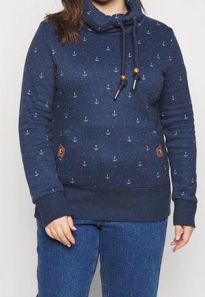 Persoon draagt een marineblauwe hoodie met witte ankerpatronen, trekkoorden met houten kralen en blauwe spijkerbroek, staand tegen een effen achtergrond.