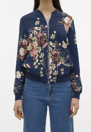 Frau trägt eine marineblaue Blumen-Bomberjacke mit Reißverschluss, gerippten Bündchen und Saum, kombiniert mit blauen Denim-Jeans.