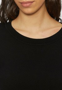 Pull noir en tricot avec un col rond côtelé, présentant une surface texturée et un matériau lisse, montrant une vue rapprochée du col.