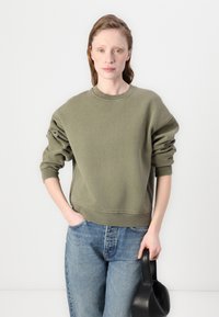 Olijfgroen oversized sweatshirt van getextureerde stof, met ronde halslijn en geribbelde manchetten, gecombineerd met lichte denim jeans en een zwarte handtas.