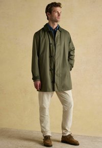 Vīrietis stāv, ģērbies olīvu zaļā mētelī, zila kreklā, bēšās biksēs un brūnās kurpēs uz bēša teksturēta fona.