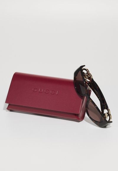 Occhiali da sole Gucci con lenti scure, montatura tartarugata e dettagli dorati, accanto a un astuccio rigido bordeaux con il nome del brand in rilievo.