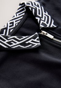 Tessuto nero con superficie strutturata, colletto a motivi geometrici bianchi e dettaglio della cerniera in argento.