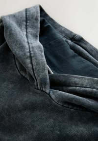 Pelēka sporta džemperis ar kapuci, kam raksturīgs teksturēts audums un brīvs apkakles dizains. Iekšpuse ir tumši zila, izceļot krāsu kontrastu.