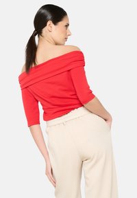 Top rosso off-shoulder con dettagli arricciati, maniche 3/4. Abbinato a pantaloni cremosi a vestibilità rilassata. Tessuto dalla superficie liscia.