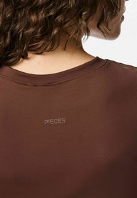 Personne aux cheveux bouclés portant une chemise brune avec le mot "PIECES" imprimé sur le haut du dos, montrée de derrière et en vue de profil.