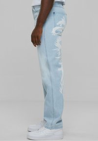 Pantalones vaqueros de mezclilla azul claro con un ajuste ligeramente holgado, que cuentan con un diseño gráfico blanco a lo largo de la pierna izquierda y se complementan con zapatillas blancas.