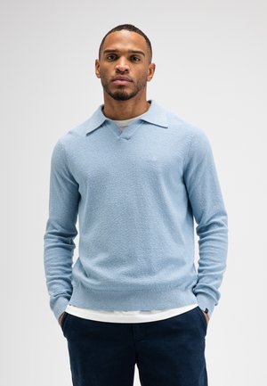 Mann trägt hellblauen strukturierten Langarmpullover über weißem Hemd mit dunkler Hose, steht mit den Händen in den Taschen vor einfarbigem Hintergrund.