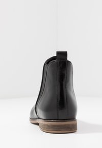Botín negro de cuero hasta el tobillo con paneles laterales elásticos, punta redonda y un tacón apilado de estilo madera. Textura suave con detalles de diseño mínimos.
