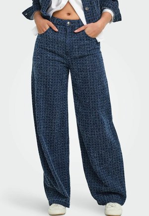 Femme portant un pantalon bleu texturé taille haute à jambes larges, les mains dans les poches, associé à des chaussures blanches et un top court.