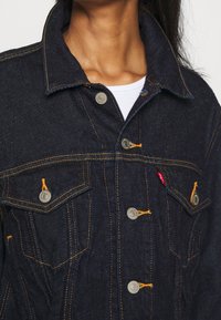 Tmavá džínová bunda se žlutým šitím, stříbrnými knoflíky a dvěma kapsami na hrudi. Má špičatý límec a volný střih.