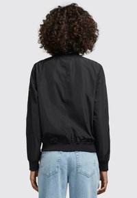 Černá bomber bunda s žebrovanými manžetami a spodním lemem, charakterizována hladkým, lesklým povrchem. Pohled zezadu ukazuje jemný branding v oblasti krku. Viditelné džíny.