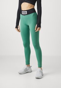 Leggings verdi con una fascia nera in vita e un'etichetta con logo, realizzati in materiale liscio e elastico, abbinati a scarpe da ginnastica bianche.
