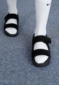 Sorte sandaler med to justerbare borrelåsremmer, åpen tå-design, båret over hvite sokker, på en blå teppeoverflate.