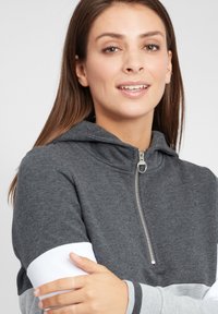 Felpa grigia con zip, dotata di cappuccio, cerniera silver e pannelli delle maniche bianchi e grigi in contrasto. Tessuto morbido con una vestibilità rilassata.
