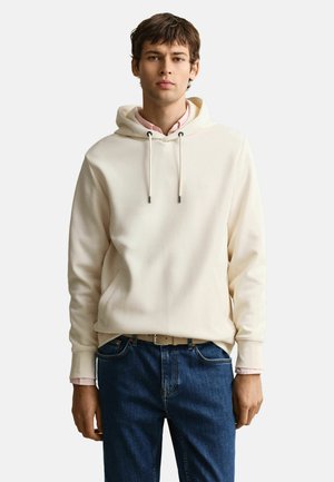 Jeune homme portant un sweat à capuche crème sur une chemise rose et un jean bleu, debout devant un fond clair uni.