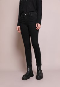 Jean skinny noirs à taille haute, en jean. Associés à des bottines noires et à un pull noir texturé, mettant en valeur une silhouette élégante.