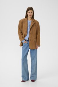 Woman wearing a brown oversized blazer, light blue buttoned cardigan, wide-leg jeans, and burgundy shoes, standing with one hand in pocket.