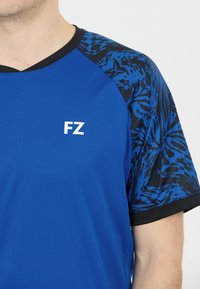 Blå sportskjorta med mönstrade ärmar i svart och blått, ribbad krage och en vit "FZ"-logotyp på framsidan. Slät textur.
